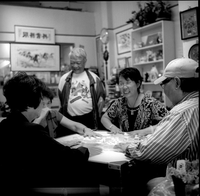 Five people gathered around a table playing a game, with a man standing and watching. Shelves and artwork decorate the background.