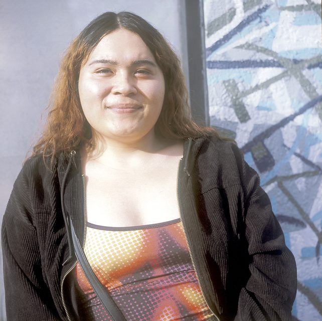 Person with long hair wearing a patterned top and black jacket stands in front of an abstract mural.
