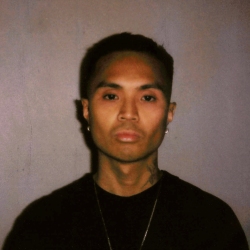 A person with short hair and earrings, wearing a black shirt, stands against a plain background.