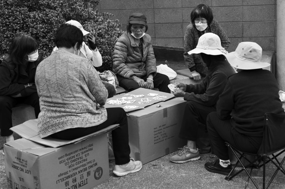 A group of women sits outdoors on cardboard boxes and chairs, playing a card game. Some wear hats and masks.