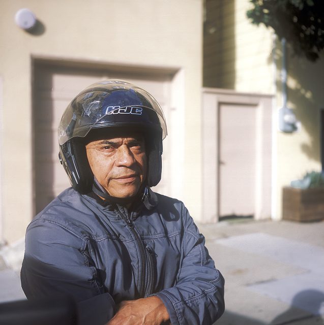 A person wearing a helmet and jacket is standing with crossed arms in front of a beige building with a garage door.
