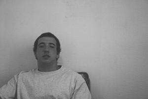 A person in a plain T-shirt sits against a blank wall, seeming relaxed and looking directly at the camera. The image is in black and white.