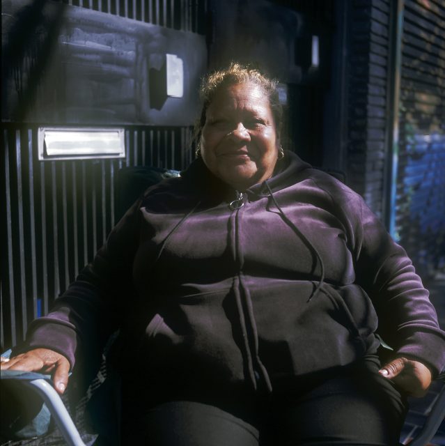 A person in a dark hoodie is sitting outdoors on a chair, with a blurred background of a building and sunlight casting shadows.