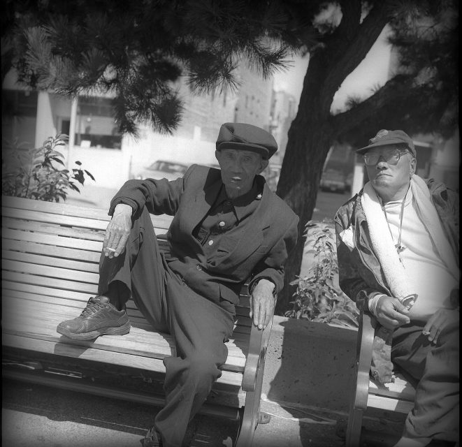 Two men sitting on a bench under a tree, one wearing a cap and blazer with crossed legs, the other in sunglasses and a cap, surrounded by an urban background.