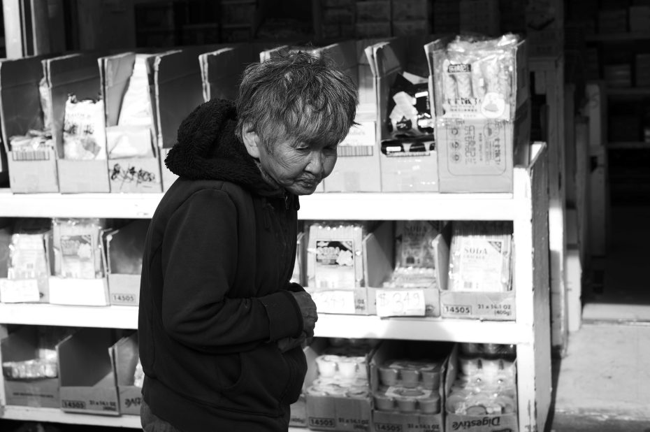 Elderly person in a hooded jacket stands near shelves with boxed goods in a store, looking down. Image in black and white.