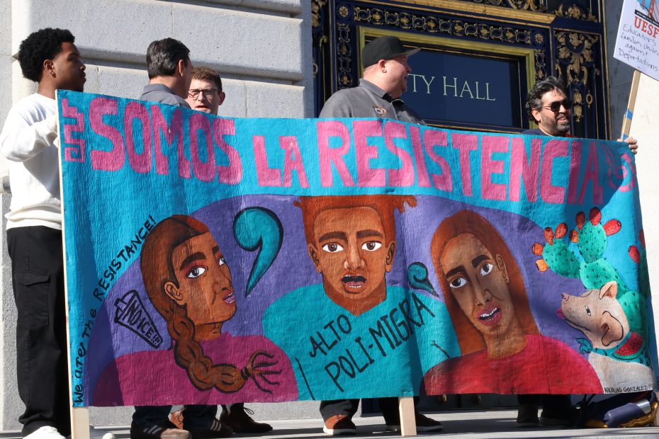 People standing outside a building hold a colorful banner with "SOMOS LA RESISTENCIA" and illustrations of faces and messages opposing deportation and ICE.