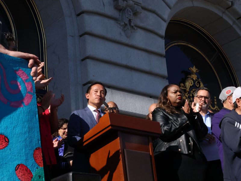 A person stands at a podium speaking to a crowd outside a building with arched windows. Others stand nearby, some clapping.