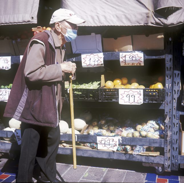 An elderly person wearing a mask and cap with a cane is walking past a fruit stand with various produce marked with prices.