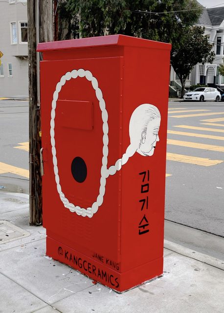 Red utility box on a street corner, featuring an artwork with a connected pattern resembling pearls, a face, and Korean text. Nearby are the names "Jane Kang" and "@KangCeramics.