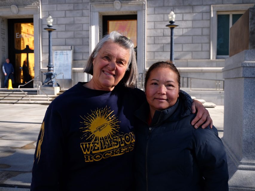 Two people stand outdoors in front of a stone building. The person on the left wears a Wellston Rockets shirt. Both are smiling and arm in arm.