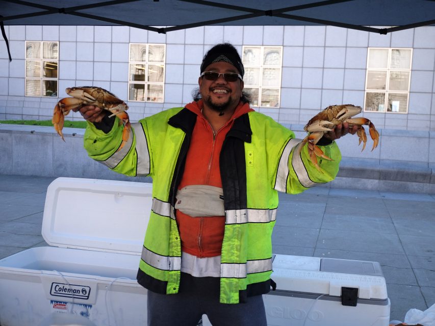 A person in a high-visibility jacket holds two crabs in front of a cooler, standing outside a building.