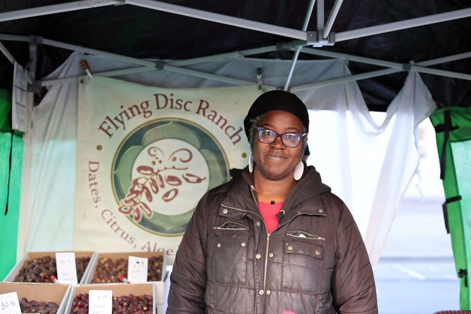 A person stands under a market tent with a sign that reads "Flying Disc Ranch." Boxes of dates are visible nearby.