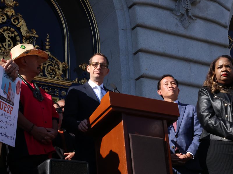 A group of people stands on the steps of a building with ornate doors, some speaking at a podium, and one holding a sign that reads "UESF Educators & Families United Against Deportations.