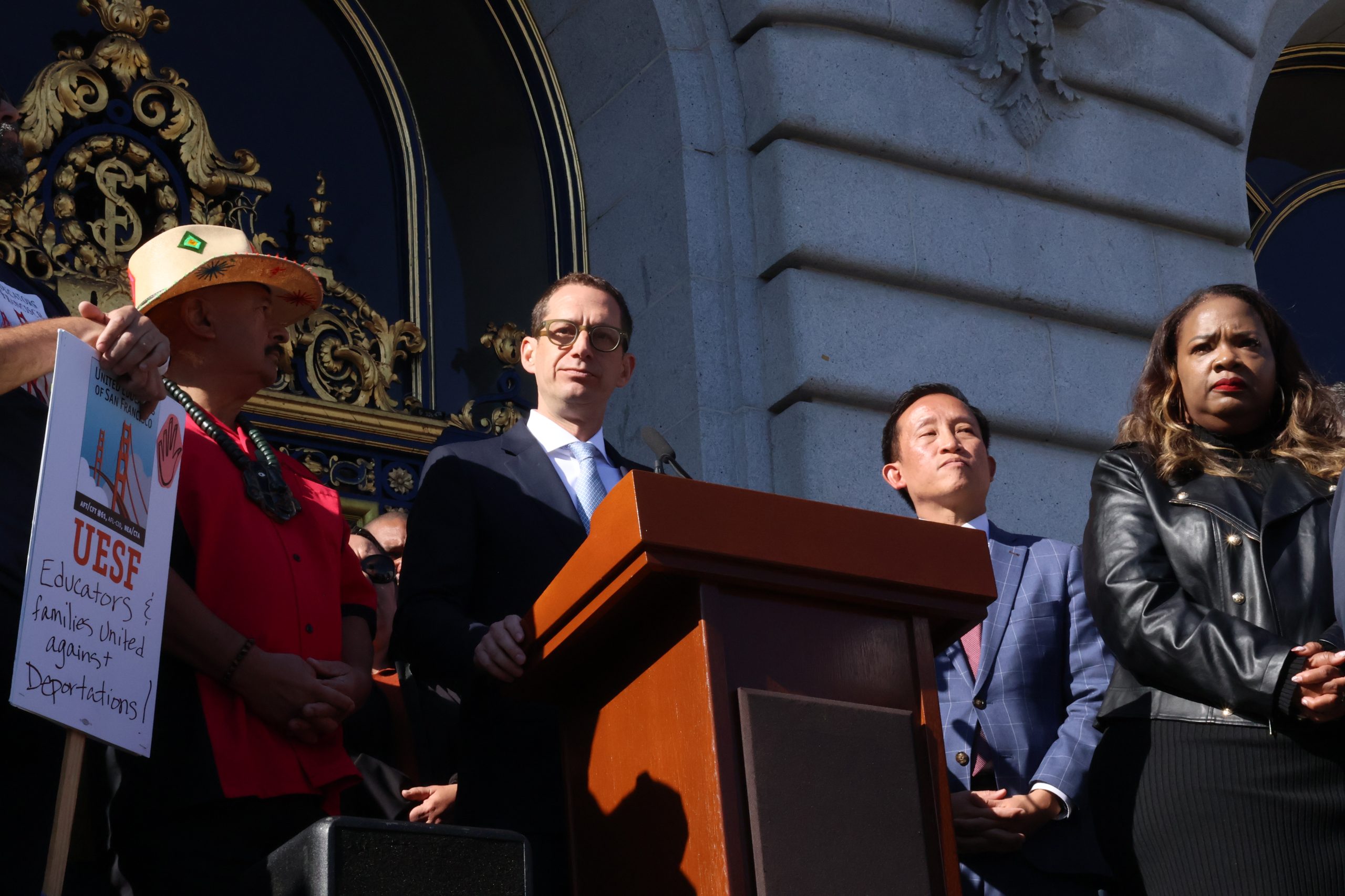 A group of people stands on the steps of a building with ornate doors, some speaking at a podium, and one holding a sign that reads "UESF Educators & Families United Against Deportations.