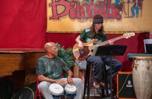 A musician sits playing conga drums, while another stands playing an electric bass guitar, next to a piano and congas, beneath a "Baobab" sign.