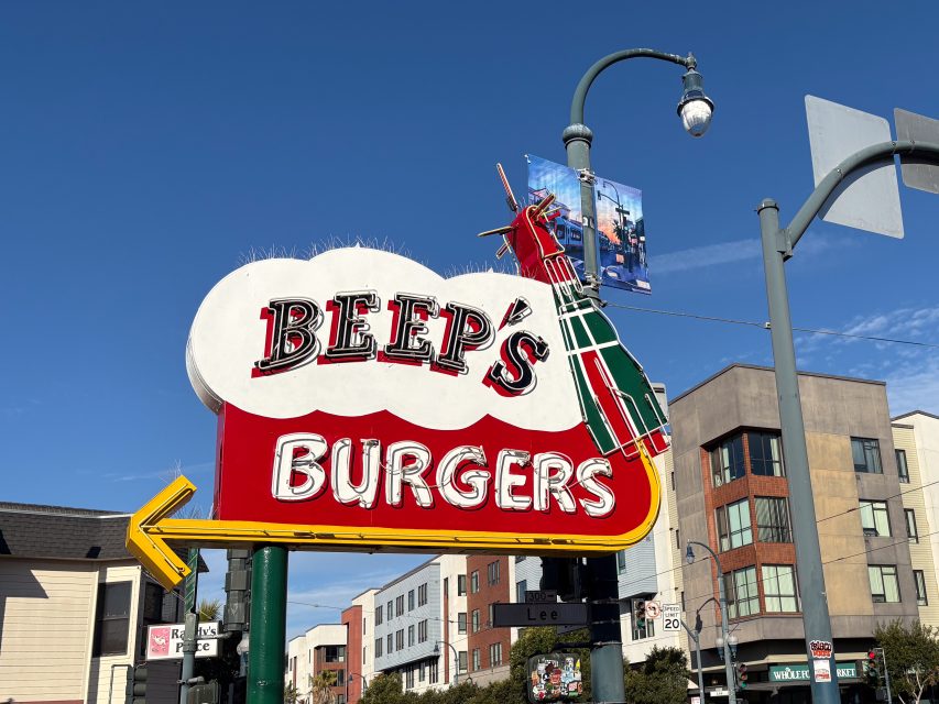 A colorful sign for Beep's Burgers featuring a cartoon character, against a clear blue sky with buildings and a streetlight in the background.