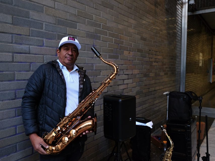 A man stands against a brick wall holding a saxophone. He is wearing a jacket and a white cap. Music equipment is visible beside him.