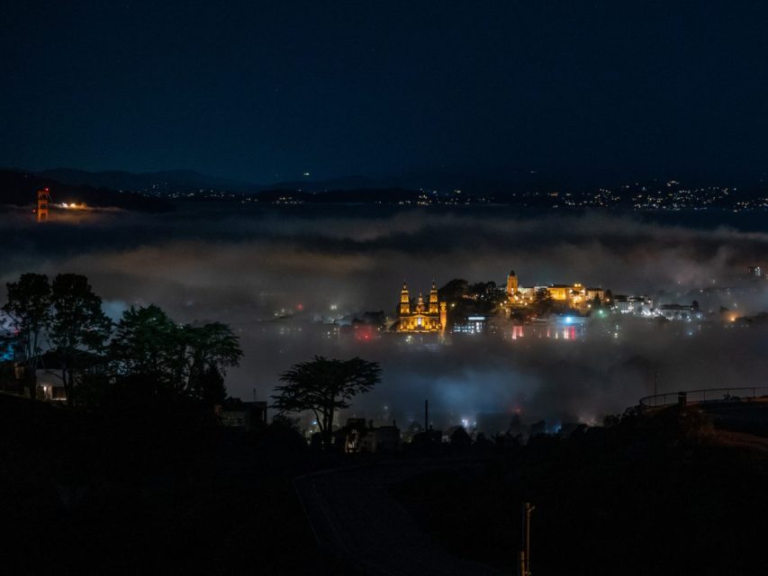 A foggy night scene with illuminated buildings and lights reflecting on water, surrounded by trees and distant hills.