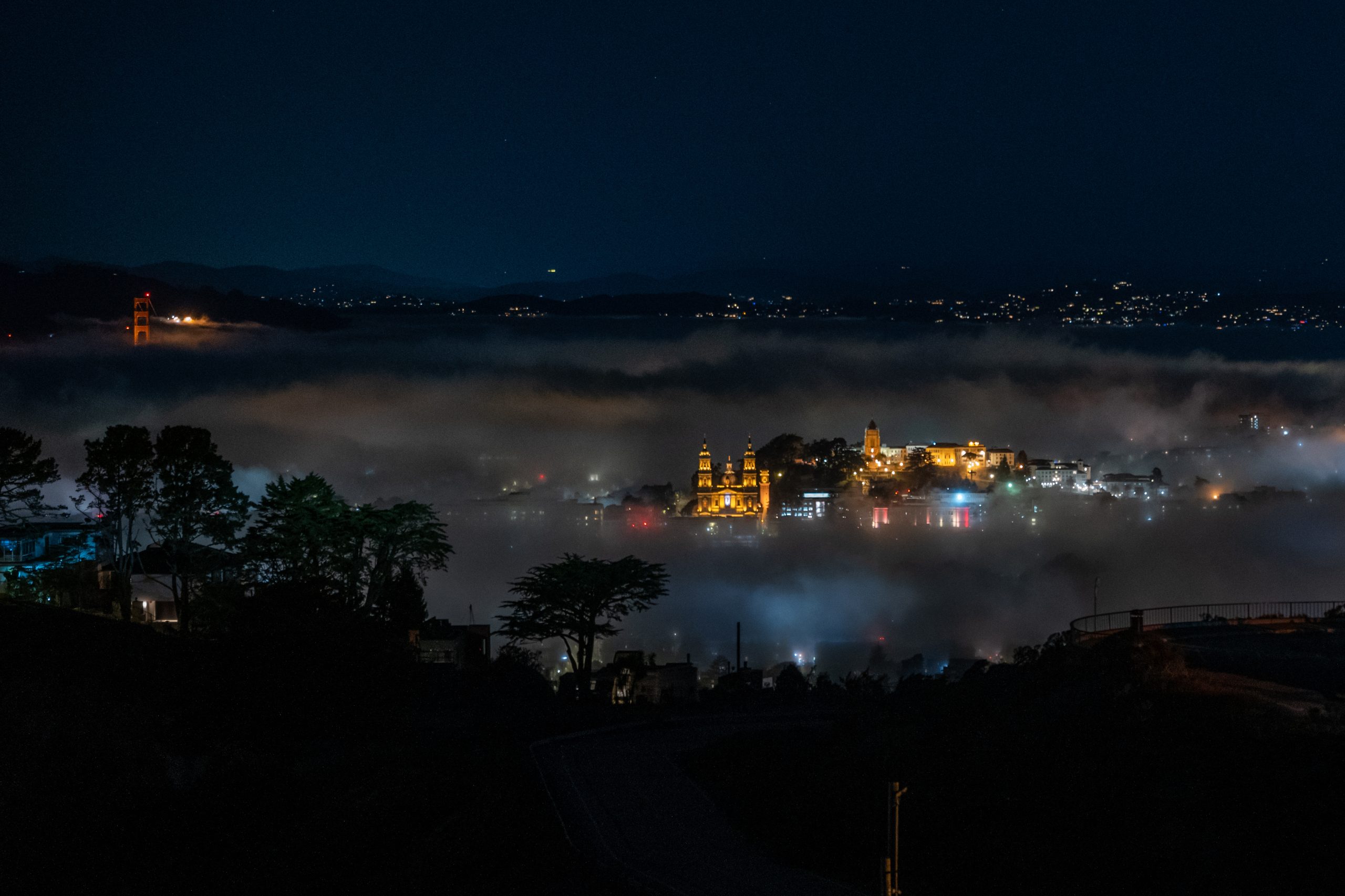 A town with illuminated buildings is partially covered in fog at night, with surrounding hills and scattered lights in the distance.