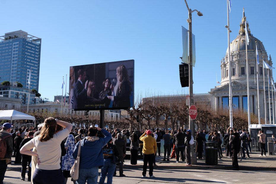 People gather outdoors in front of a large screen displaying a group of individuals. Buildings and a dome structure are visible in the background.