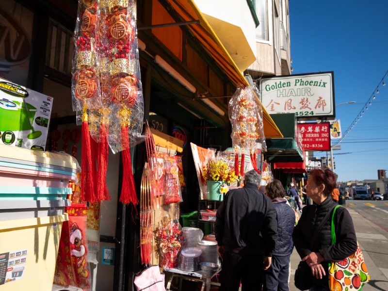 People walk by a storefront displaying red and gold decorations in a Chinatown area. A sign for Golden Phoenix Hair is visible.