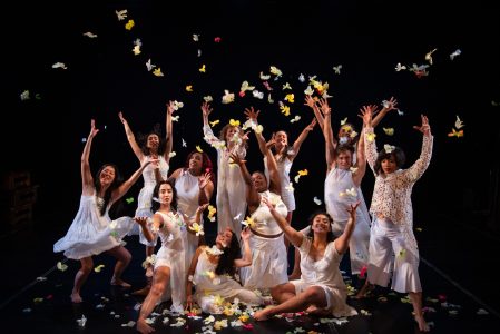 A group of individuals in white clothing joyfully throw colorful petals or butterflies in the air on a dark stage.