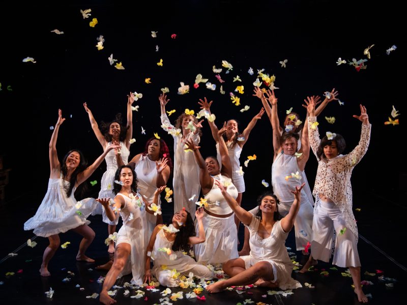 A group of individuals in white clothing joyfully throw colorful petals or butterflies in the air on a dark stage.