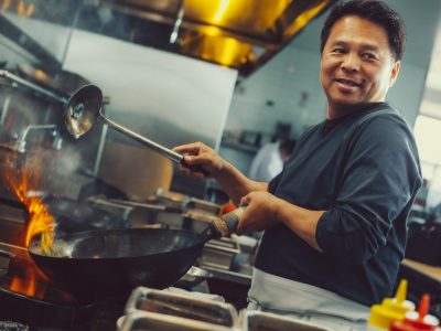 Chef cooking with a wok over a flame in a commercial kitchen, holding a ladle and smiling.