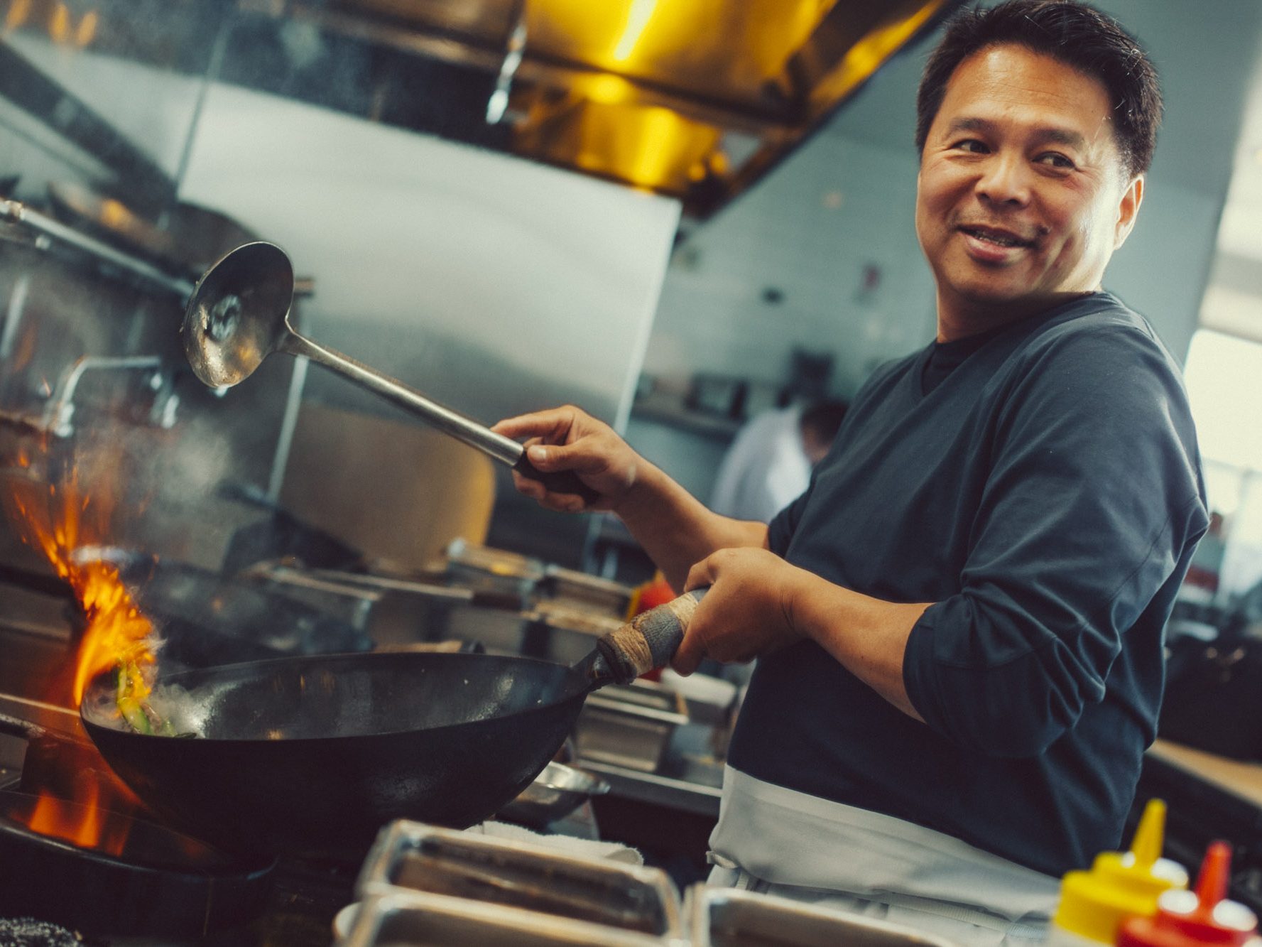 Chef cooking with a wok over a flame in a commercial kitchen, holding a ladle and smiling.