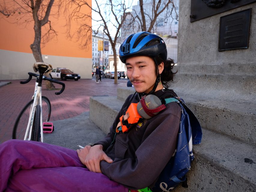 Person wearing a helmet and sitting on concrete steps beside a bicycle in an urban setting.