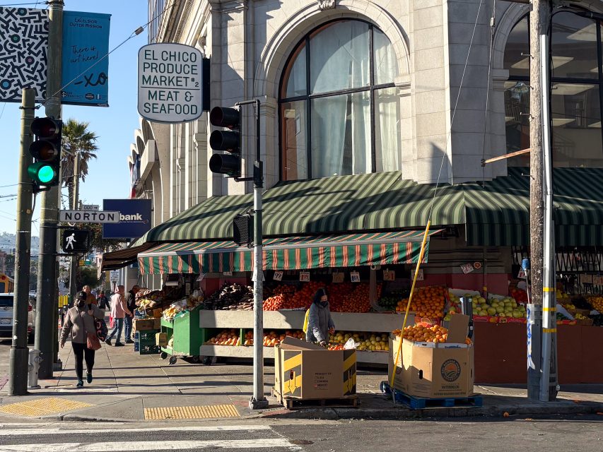 Street corner with a produce market displaying fruits and vegetables under a green awning. Traffic lights and signs are visible. A few people walk by, and cardboard boxes are on the sidewalk.