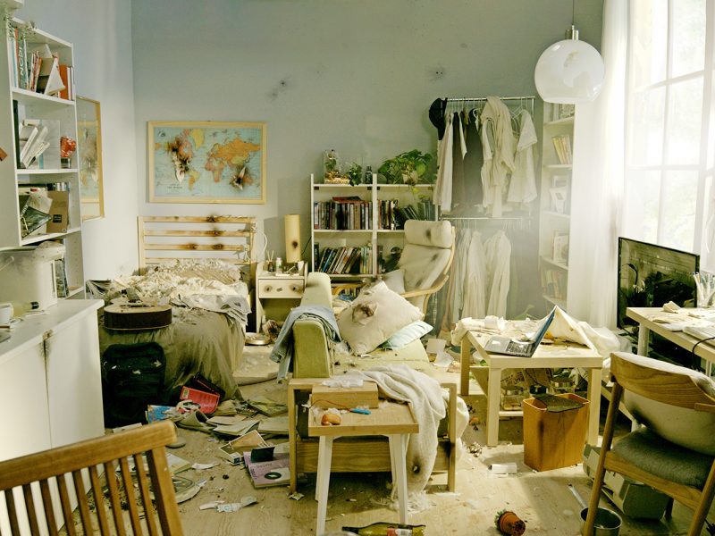 A cluttered bedroom with an unmade bed, scattered clothes, and books resembles a miniature museum of daily life. A laptop on the desk waits amid organized chaos. Shelves display various items as if curated for curious onlookers, while sunlight streams through a large window, illuminating the space.
