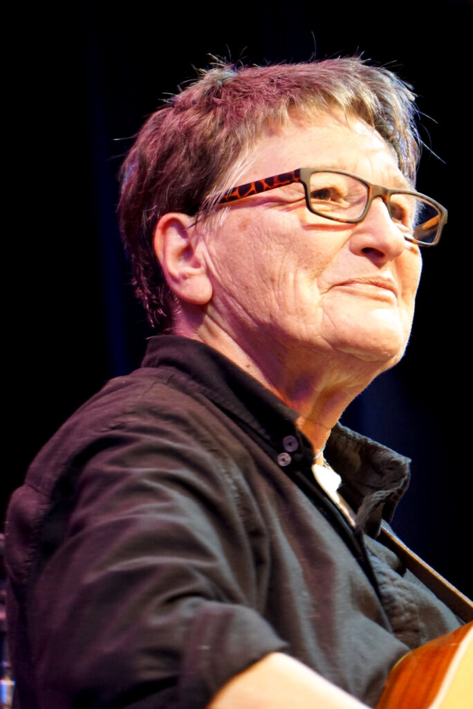 Person with short hair and glasses in a black shirt, looking to the side against a dark background.