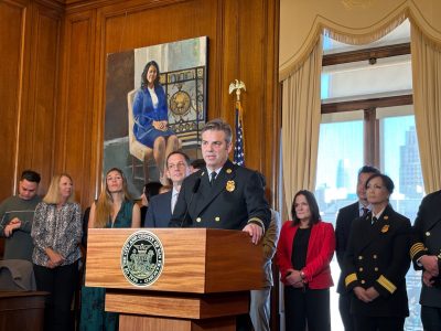 A uniformed official speaks at a podium with several people standing behind in a room, a painting and American flag in the background.