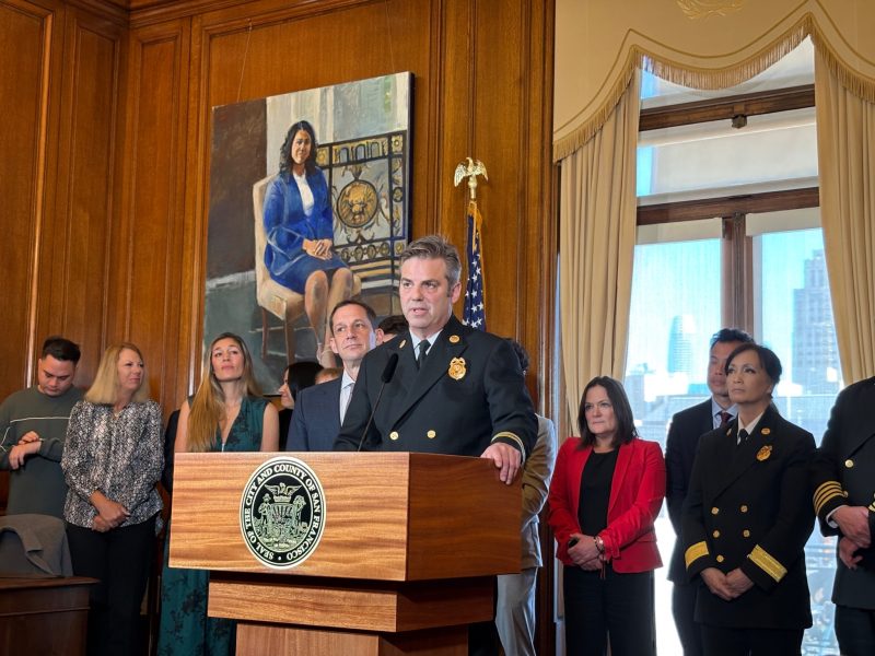 A uniformed official speaks at a podium with several people standing behind in a room, a painting and American flag in the background.