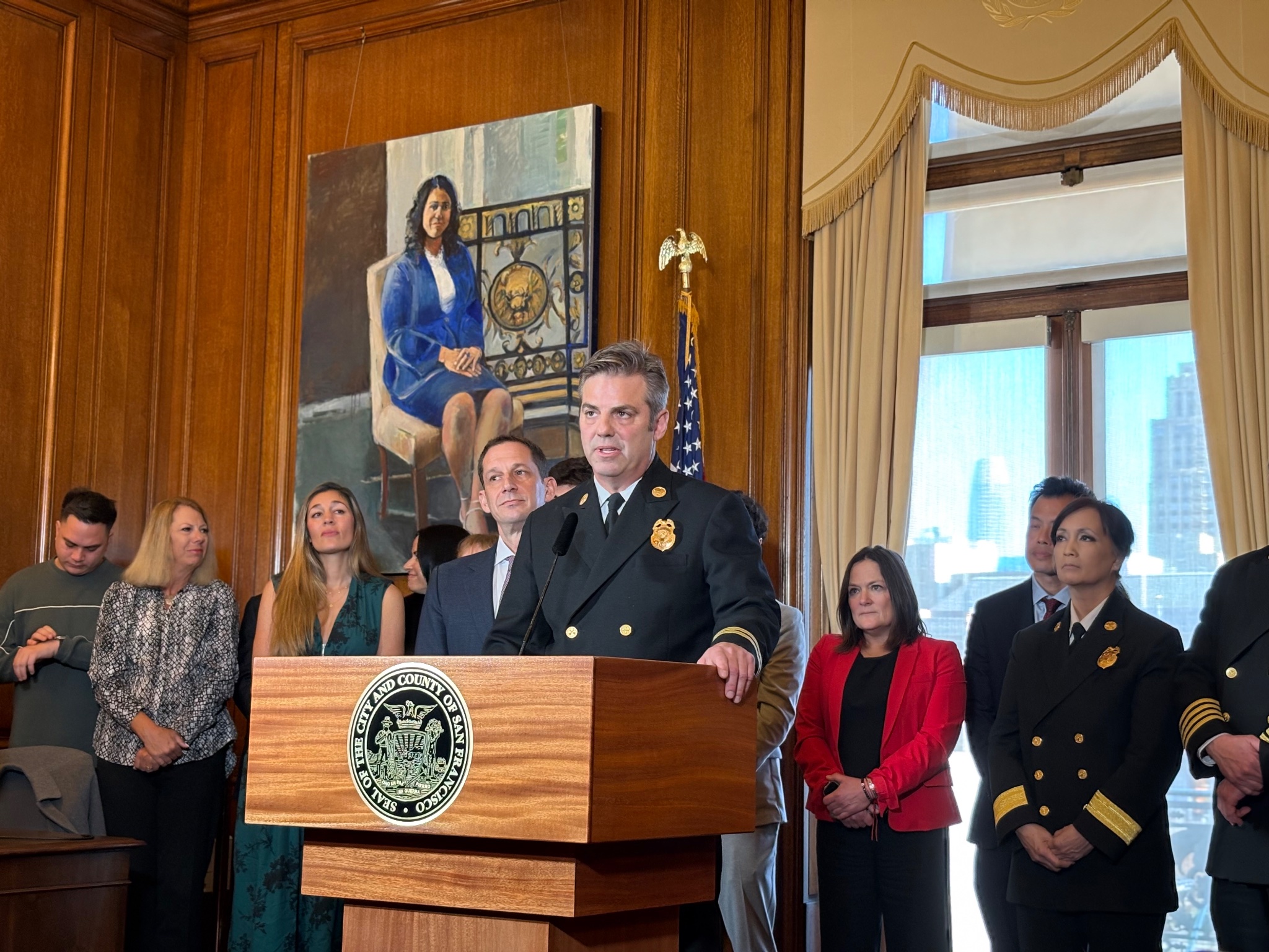 A uniformed official speaks at a podium with several people standing behind in a room, a painting and American flag in the background.