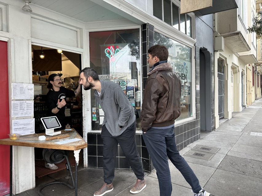 Two men stand outside a street-front cafe, engaging in conversation with a staff member inside. A small wooden table with a payment terminal is in front of the entrance.