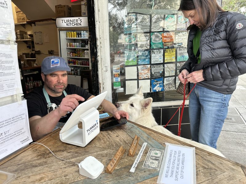 A man operates a touchscreen register at Turner's Kitchen. A white dog on a leash sits near the counter while a woman stands nearby holding the leash.