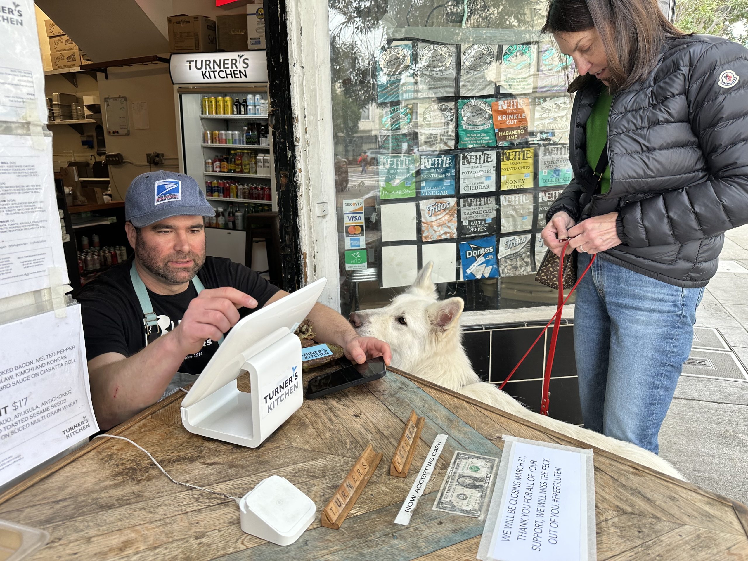 A man operates a touchscreen register at Turner's Kitchen. A white dog on a leash sits near the counter while a woman stands nearby holding the leash.