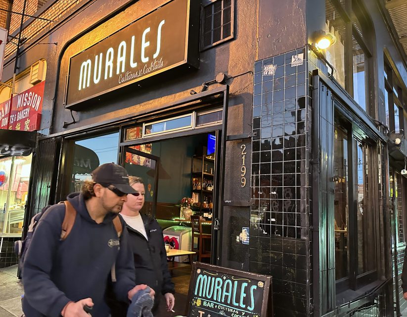 A man and woman stroll past Murales Bar and Kotails, a black-tiled establishment with a glowing interior. The entrance proudly displays the street number 2199, inviting passersby to discover its vibrant ambiance.