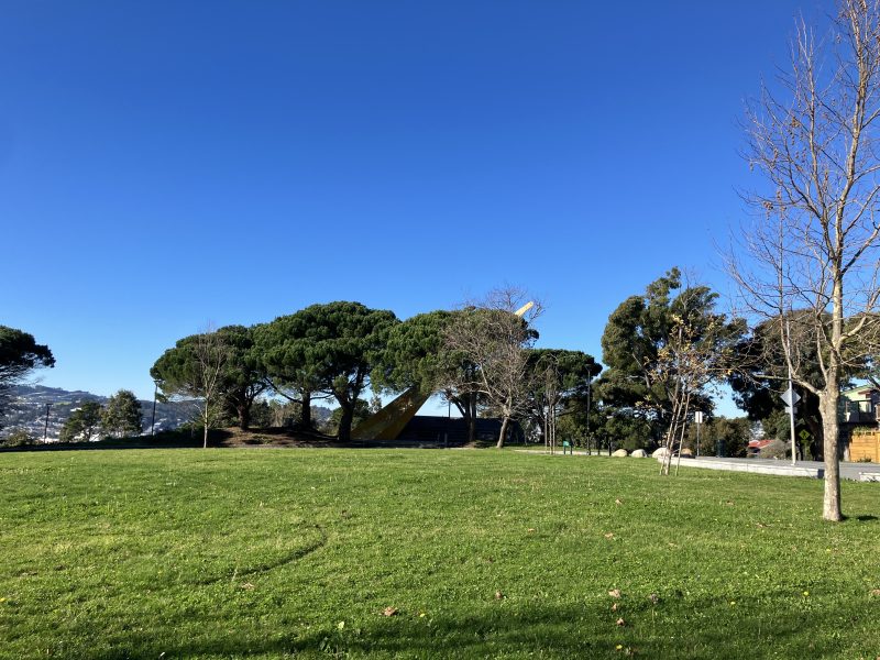 A clear blue sky over a grassy field with a row of green and bare trees. A large yellow sculpture is visible among the trees.