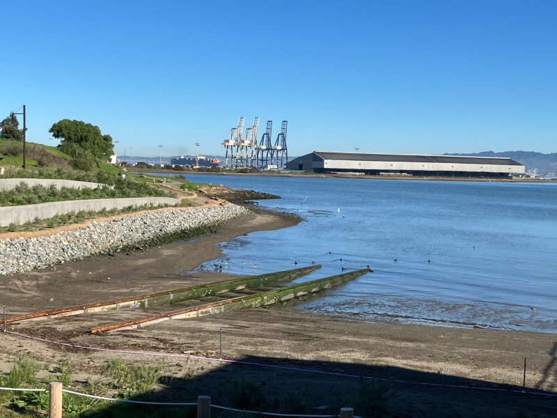 View of a coastal area with a small boat ramp in the foreground, cranes, and an industrial building in the background, on a clear day.