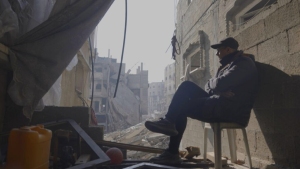 A person in a cap sits on a stool amidst a building's rubble, looking out at a street with damaged structures in daylight.