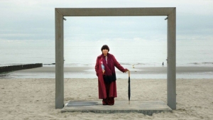 Person in a long coat holding an umbrella stands on a platform framed by a large square structure on a sandy beach. The sea and cloudy sky are in the background.