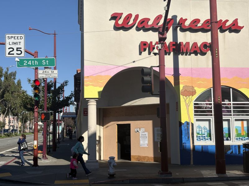 Street scene at an intersection with a Walgreens pharmacy and a mural on the building. A person and a child are walking near the corner under a red traffic light.