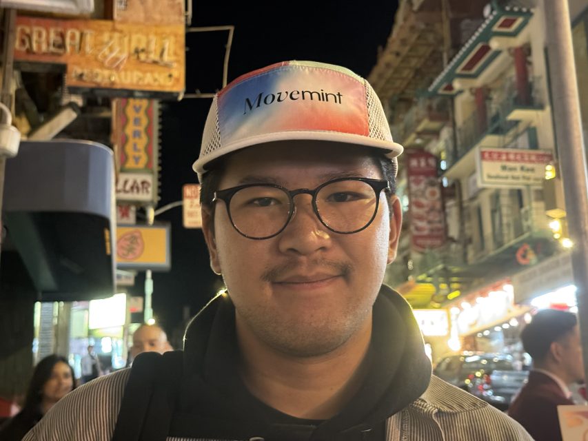 A person wearing glasses and a multicolored hat stands on a bustling city street at night, with neon signs and buildings in the background.