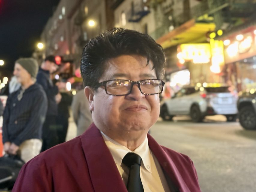 A person wearing glasses and a maroon jacket stands on a busy city street at night, with people and cars in the background.