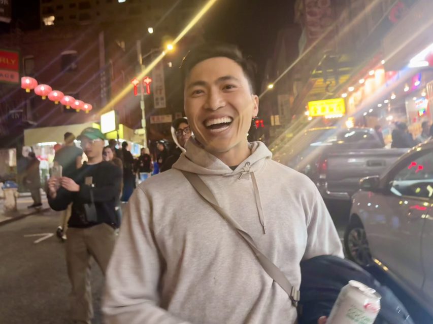 Quan Pham in a gray hoodie smiling on a busy, brightly-lit city street at night, holding a can and carrying a bag. Pham voted for Lurie.