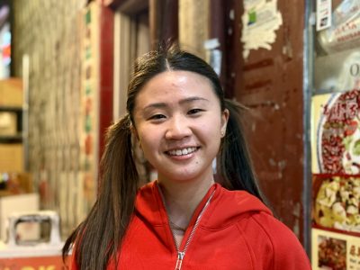 A person in a red hoodie smiles while standing in front of a wall with various posters and signs.