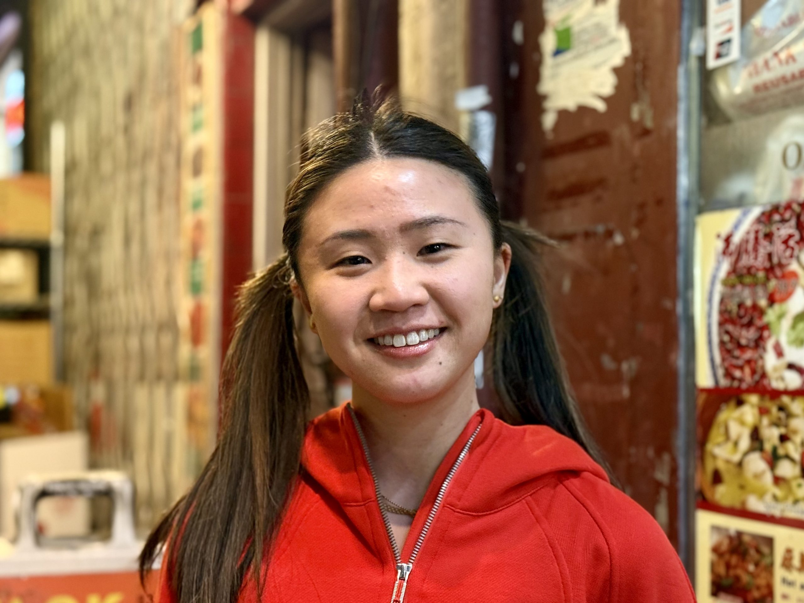 A person in a red hoodie smiles while standing in front of a wall with various posters and signs.
